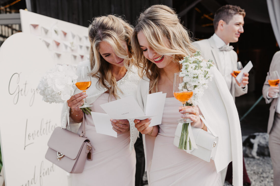 Zwei lachende Hochzeitsgäste in pastellfarbenen Kleidern lesen ihre persönlichen Nachrichten beim Sektempfang.