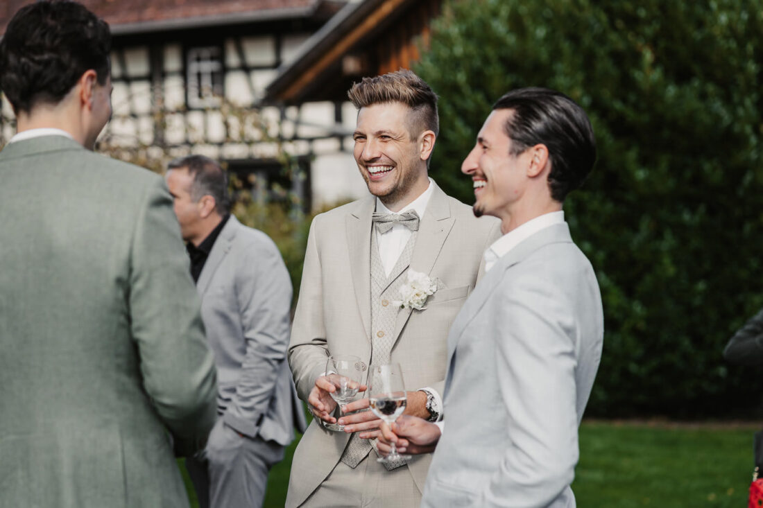 Drei elegant gekleidete Männer lachen bei einem Sektempfang im Freien auf einer Hochzeit im Steinbachhof, im Hintergrund ein historisches Fachwerkhaus.