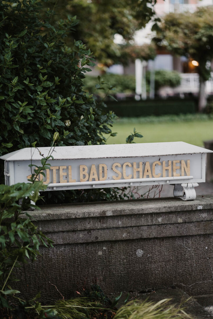 Goldener Schriftzug "Hotel Bad Schachen" auf einer weißen Steinmauer im Garten der Location in Lindau.