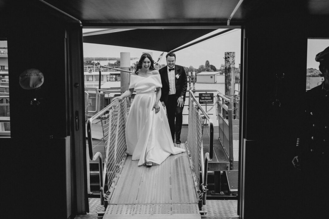 Schwarz-Weiß-Foto eines glücklichen Brautpaares, das gemeinsam über den Metallsteg auf das Schiff zuläuft.
