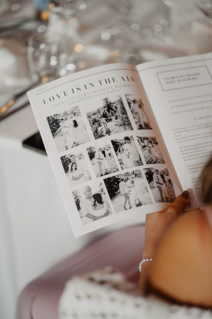 Ein Gast hält eine aufgeschlagene Hochzeitszeitung in der Hand mit Schwarz-Weiß-Fotos des Brautpaares.