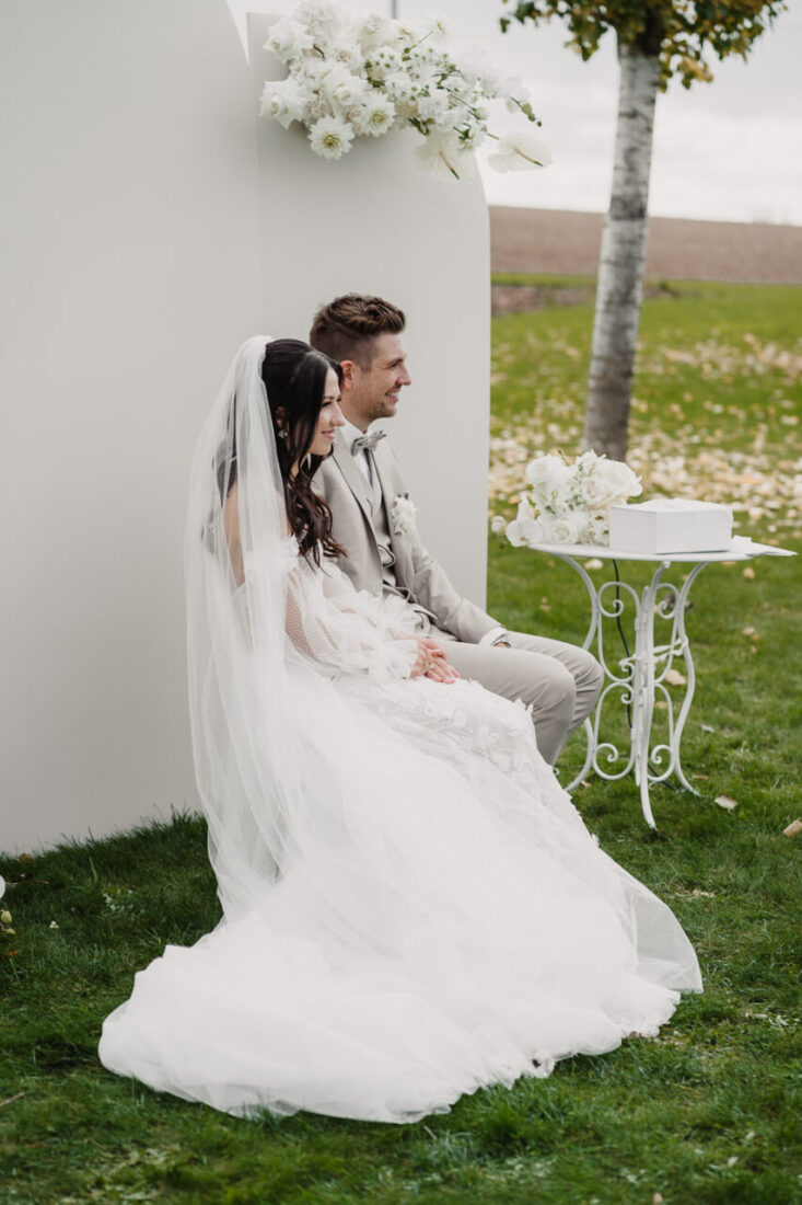 Braut und Bräutigam sitzen während der freien Trauung vor weißer Blumendeko auf dem Steinbachhof.