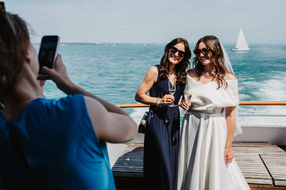 Die Braut und ihre Freundin stoßen mit Sonnenbrillen und einem Glas Sekt auf dem Sonnendeck des Schiffes an.
