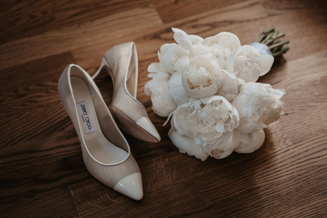 Elegante Jimmy Choo Brautschuhe und ein weißer Pfingstrosen-Brautstrauß auf Holzboden fotografiert.