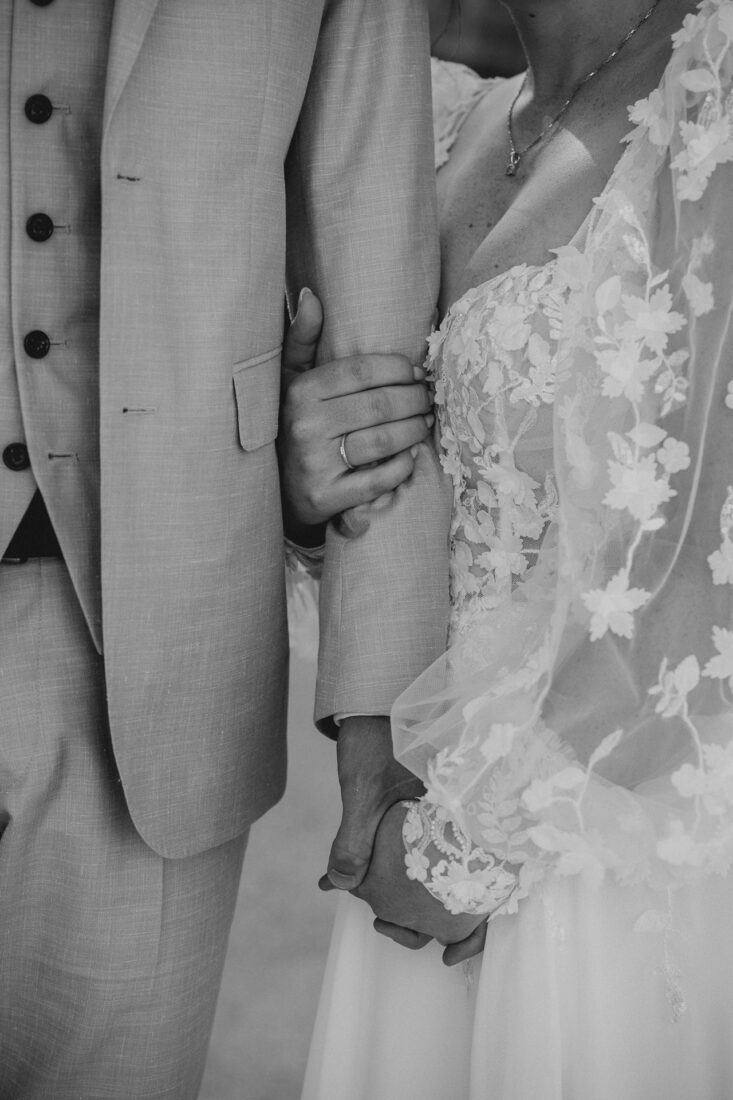 Detailaufnahme der Eheringe, während die Braut den Arm des Bräutigams umschlingt.