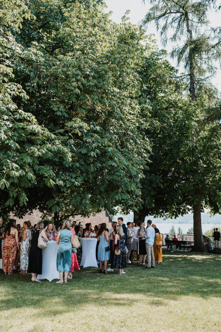 Viele Hochzeitsgäste stehen bei sommerlichem Wetter unter großen Bäumen im Garten hinter dem Museumssaal in Überlingen mit Blick auf den Bodensee.