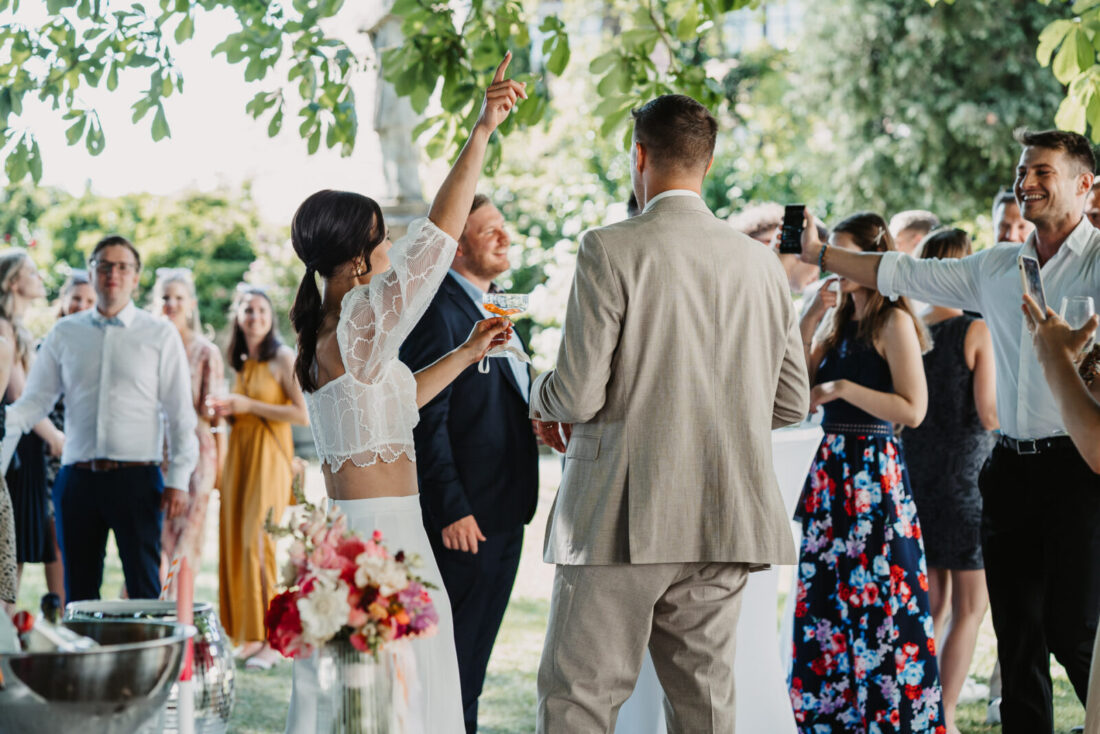 Die Braut reckt jubelnd den Arm mit einem Drink in die Höhe, umgeben von lachenden Gästen im Außenbereich des Museumssaals.