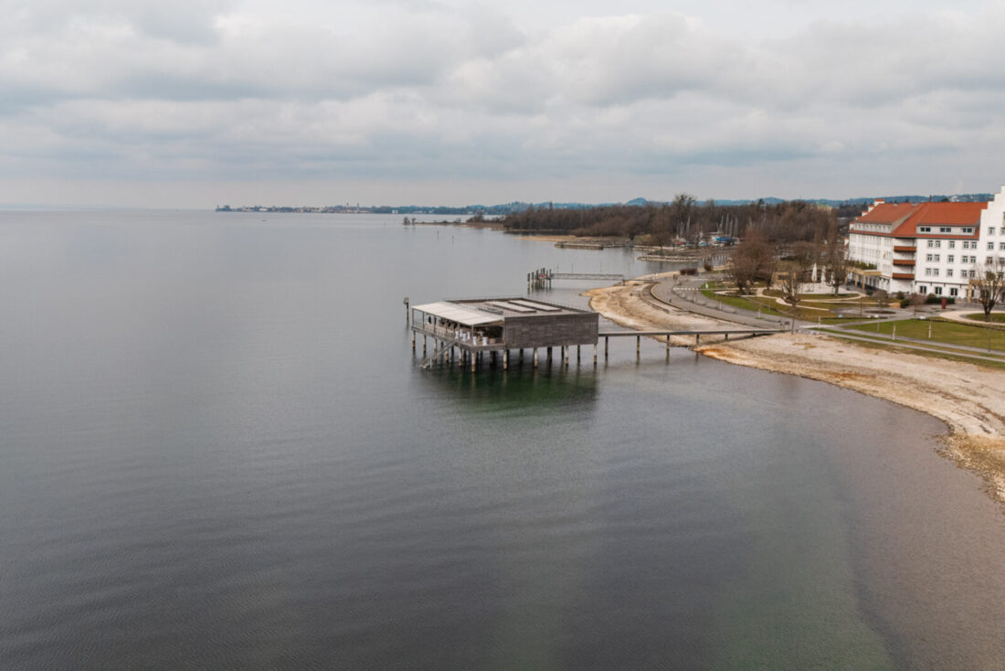 Luftaufnahme an einem regnerischen Tag vom Badehaus des Seehotels Kaiserstrand in Lochau. Man sieht das Hotel dezent im Hintergrund, während der Bodensee und die Insel von Lindau gut zu sehen sind.
