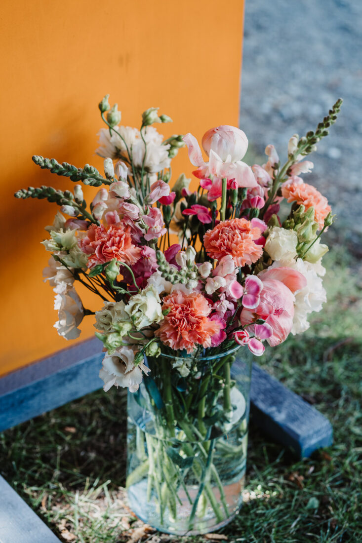 Ein farbenfroher Hochzeitsstrauß mit Nelken und Löwenmäulchen in einer Glasvase vor einer orangefarbenen Wand im Museumssaal Überlingen.
