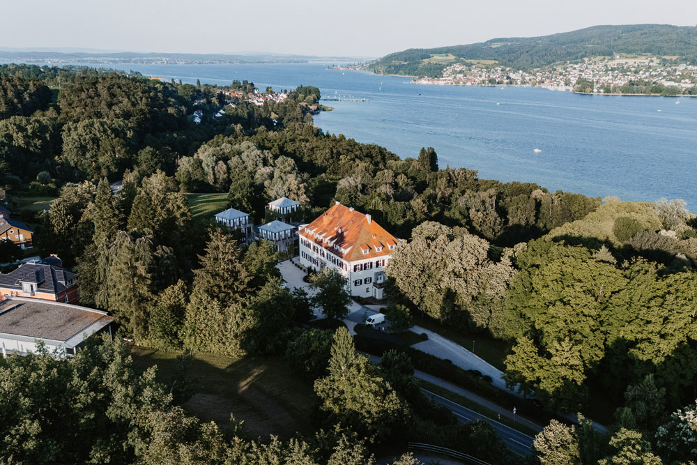 Luftaufnahme vom Schloss Marbach umgeben der grünen Parkanlage und dem Bodensee im Hintergrund.