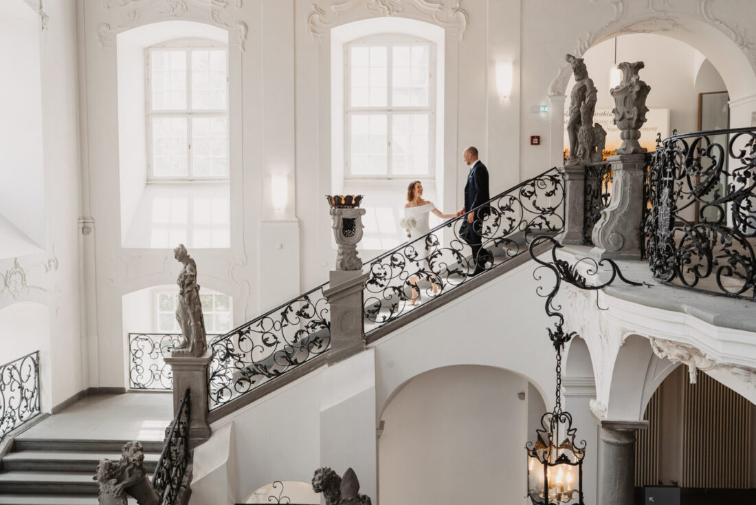Weitwinkel-Aufnahme des Brautpaares im prunkvollen, hellen Barock-Treppenhaus des Neuen Schlosses Meersburg mit historischen Statuen.