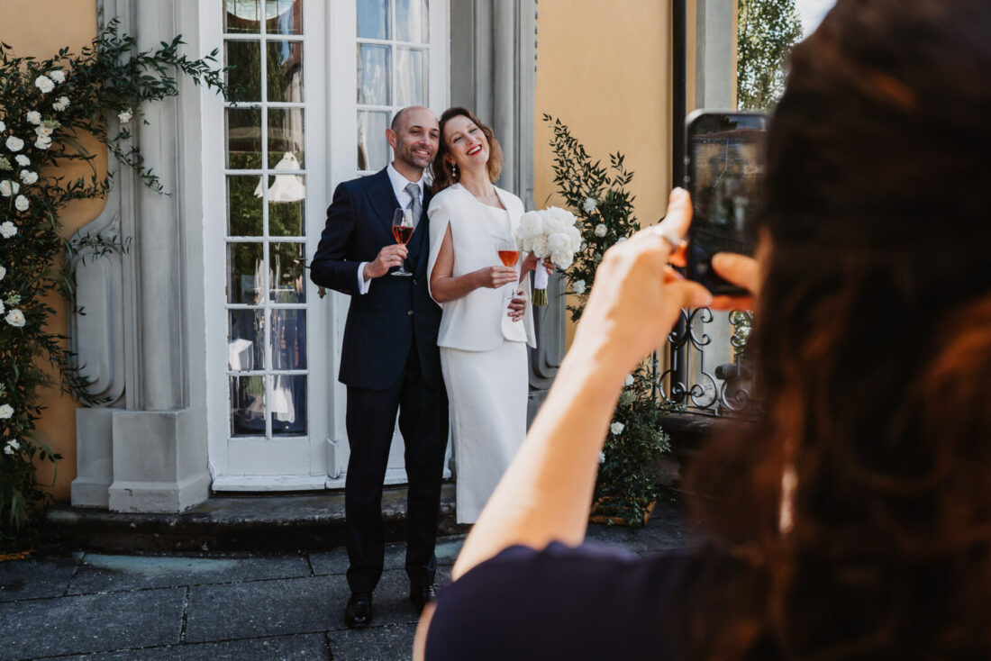 Brautpaar stößt mit Rosé-Sekt an, im Vordergrund fotografiert ein Gast den Moment mit dem Smartphone.