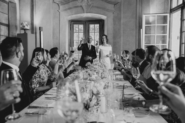 Schwarz-Weiß-Aufnahme des Brautpaares, das während einer Rede die Gläser erhebt und mit den Gästen anstößt.