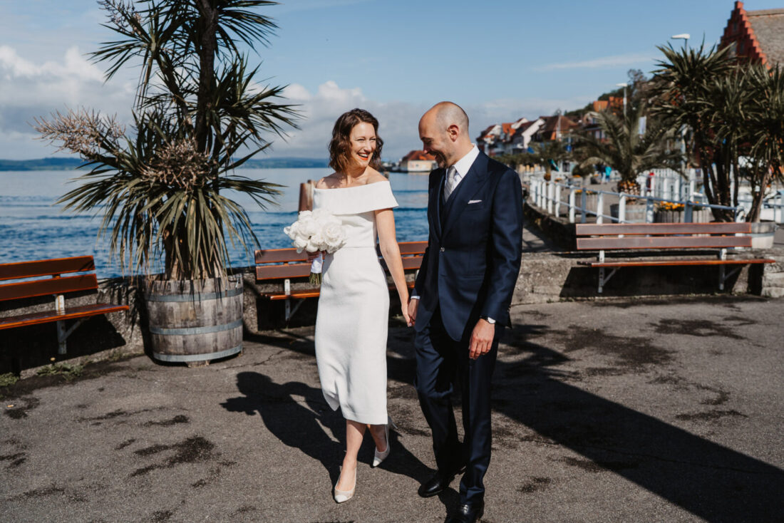 Brautpaar läuft lachend über die Promenade in Meersburg, im Hintergrund Palmen und der sommerliche Bodensee.