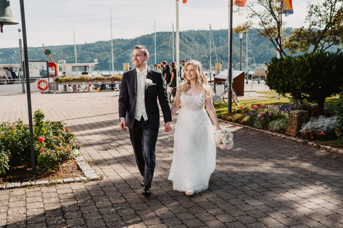 Das Brautpaar läuft Hand in Hand Richtung ihrer Feierlocation im Zollhaus in Bodman-Ludwigshafen. Im Hintergrund ist der See, das Schiff und die Hochzeitsgäste zu erkennen.