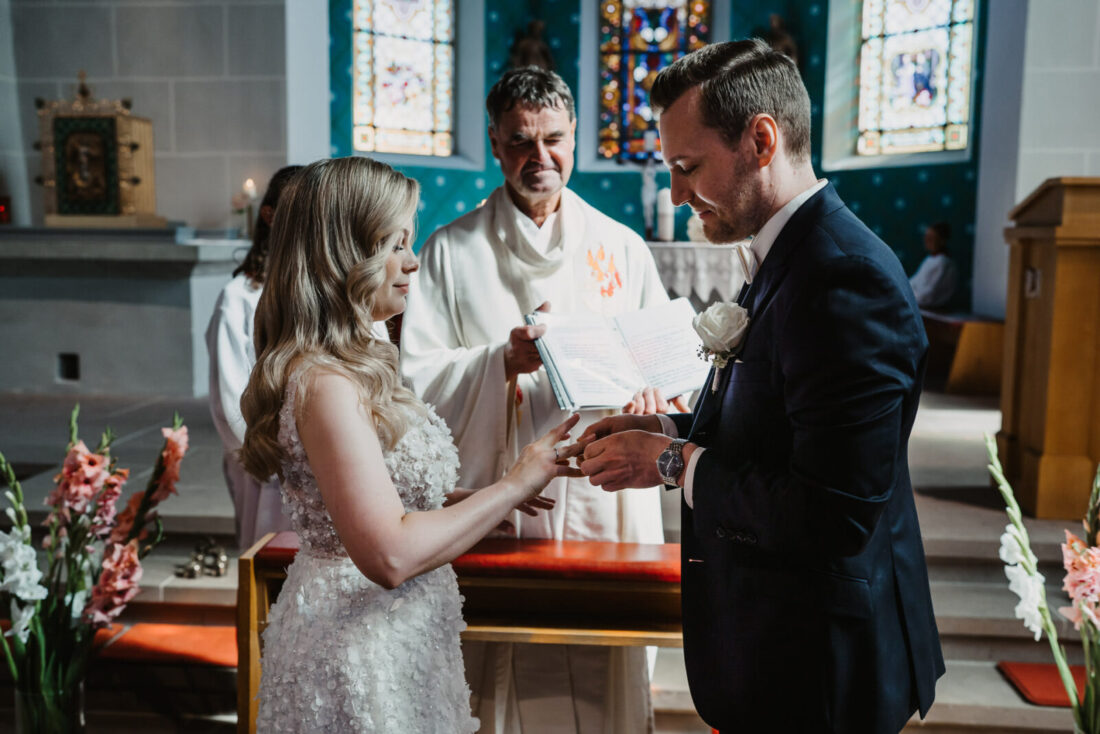 Der Bräutigam steckt seiner Braut den Ehering an, während der Pfarrer im Hintergrund das Buch mit dem Ehegelübde hält.