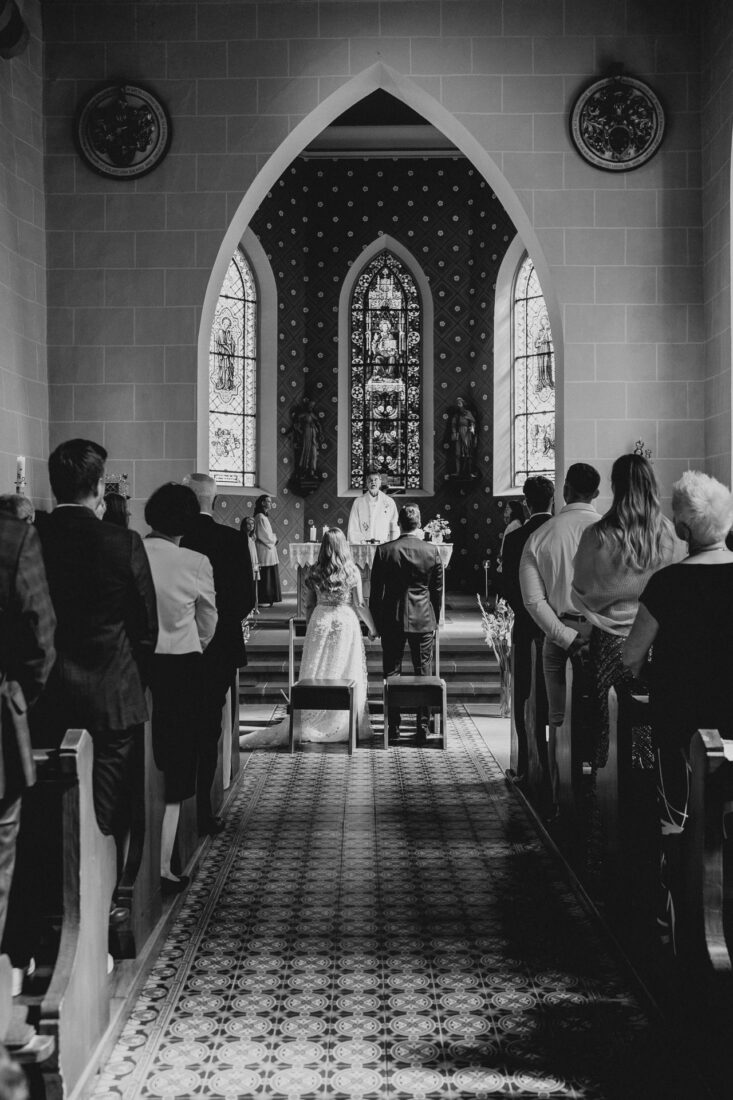 Das Brautpaar und seine Gäste stehen bei ihrer kirchlichen Trauung in Bodman-Ludwigshafen und beten. Fotografiert dezent aus dem Hintergrund am Eingang der Kirche